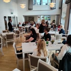 In einem hellen Raum hält ein Mann einen Vortrag vor einer aufmerksam lauschenden Gruppe, während jemand ein Foto macht.