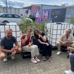 Fünf Personen sitzen an einem sonnigen Tag auf Kisten, genießen Eis und Getränke vor Gebäuden mit bunten Werbeplakaten.