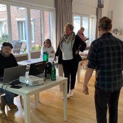 Eine Gruppe von Menschen diskutiert in einem hellen Raum mit Laptops und Getränken auf den Tischen.