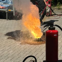 Ein Mann führt eine Feuerlöschübung durch, während ein Feuerball auf dem Boden auflodert und Feuerlöscher bereitstehen.