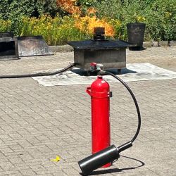 Ein roter Feuerlöscher steht bereit, während Flammen aus einer Metallkonstruktion auf dem Pflaster lodern.