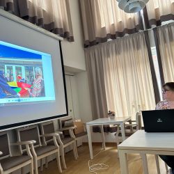 Eine Frau präsentiert einen Bildschirm mit Senioren, die mit einem bunten Tuch spielen, in einem hellen Raum.