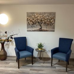Zwei blaue Sessel stehen einladend vor einem blühenden Baumgemälde, flankiert von einer Vase mit Zweigen und einem Beistelltisch.