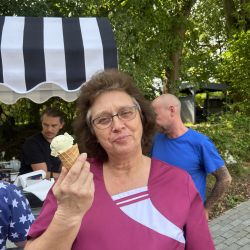 Eine Frau genießt ein Eis vor einem schwarz-weiß gestreiften Stand im Freien, umgeben von grünen Bäumen.