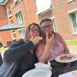 Zwei Frauen genießen Pizza auf einer sonnigen Terrasse vor einem charmanten Backsteingebäude.