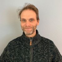 Ein lächelnder Mann in einem warmen Strickpullover blickt in die Kamera vor einem neutralen Hintergrund.