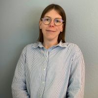 Eine Person mit Brille und gestreiftem Hemd lächelt leicht vor einer grauen Wand.