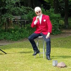 Ein Mann in einem roten Sakko sitzt auf einem Hocker und spricht draußen in ein Mikrofon, umgeben von grüner Natur.