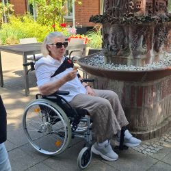 Eine ältere Frau im Rollstuhl genießt ein Eis neben einem Brunnen in einem blühenden Garten. | © Name
