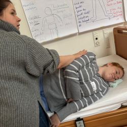 Eine Frau führt Pflegeübungen auf einem Bett durch, während Anleitungsplakate mit Zeichnungen die Wand dekorieren.