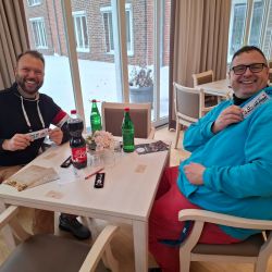 Zwei lachende Männer sitzen in einem Café und halten lächelnd Schokoriegel mit der Aufschrift "du" in die Kamera.