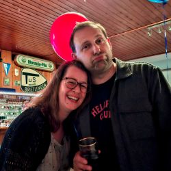 Ein fröhliches Paar posiert in einer Bar mit Luftballons, während einer einen Drink in der Hand hält. | © Name