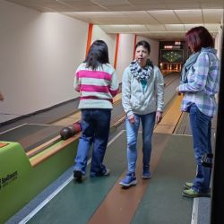 Drei Personen unterhalten sich lebhaft neben einer Kegelbahn, während eine Kugel die Bahn hinunterrollt.