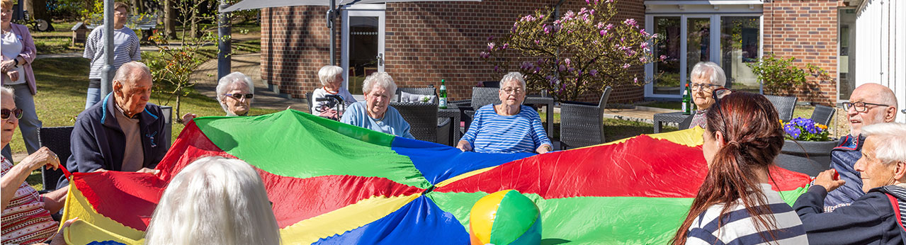Ältere Menschen spielen im Freien mit einem bunten Fallschirm und genießen eine fröhliche Veranstaltung im Sonnenschein.